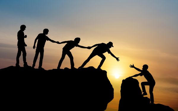 Silhouette of four people joining hands to help a person climb up a mountain, set against a vibrant sunset sky.