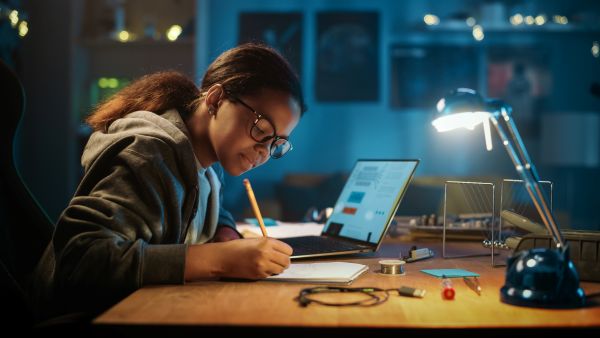 Female teen sitting at a desk writing notes and researching on computer