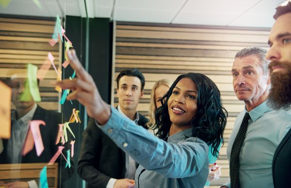 Female professional pointing at sticky notes on a board in front of a group of coworkers