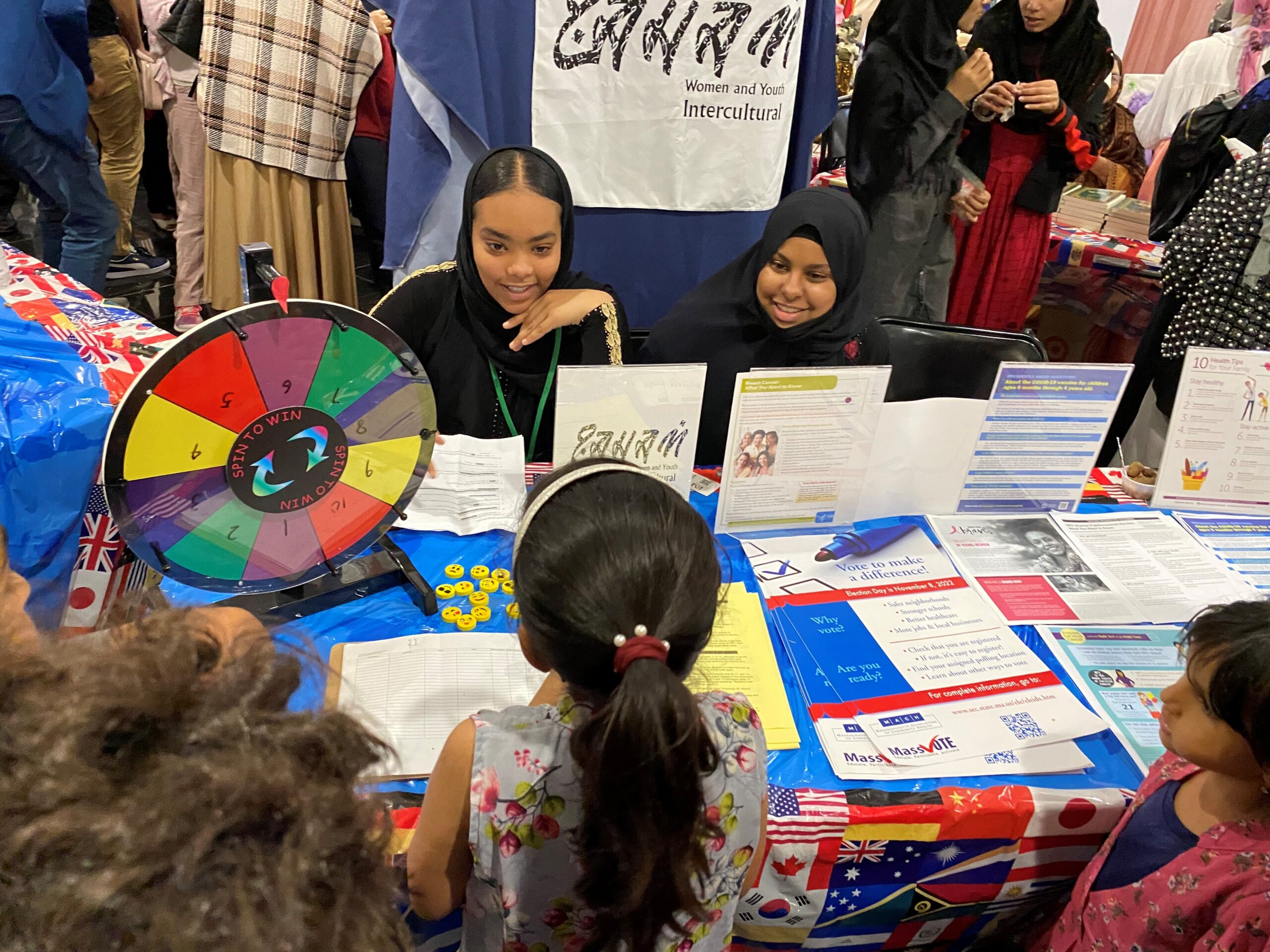 two young women speaking to young children at a table during a community event