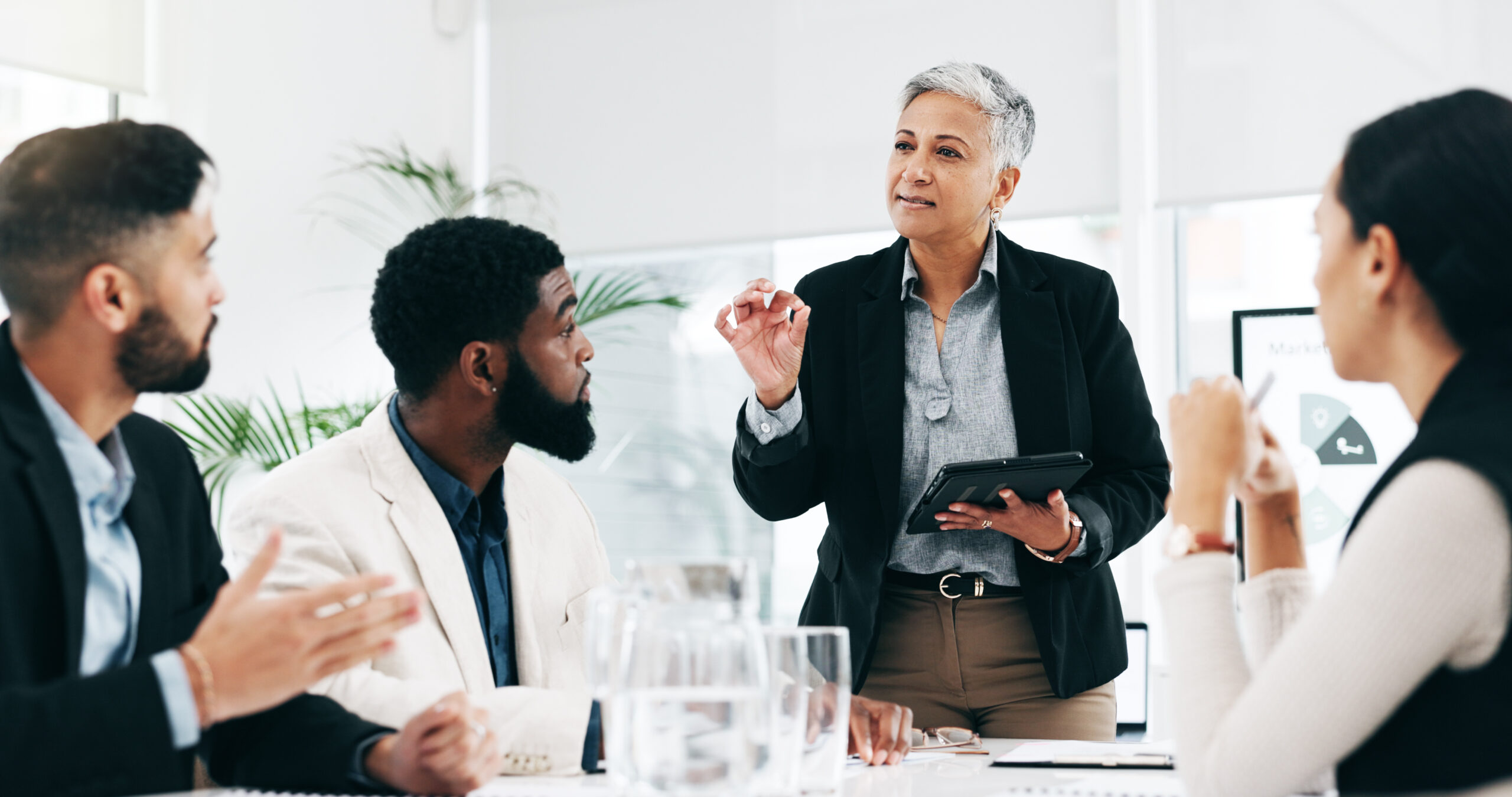 a business woman holding a tablet speaking to other business people at a table