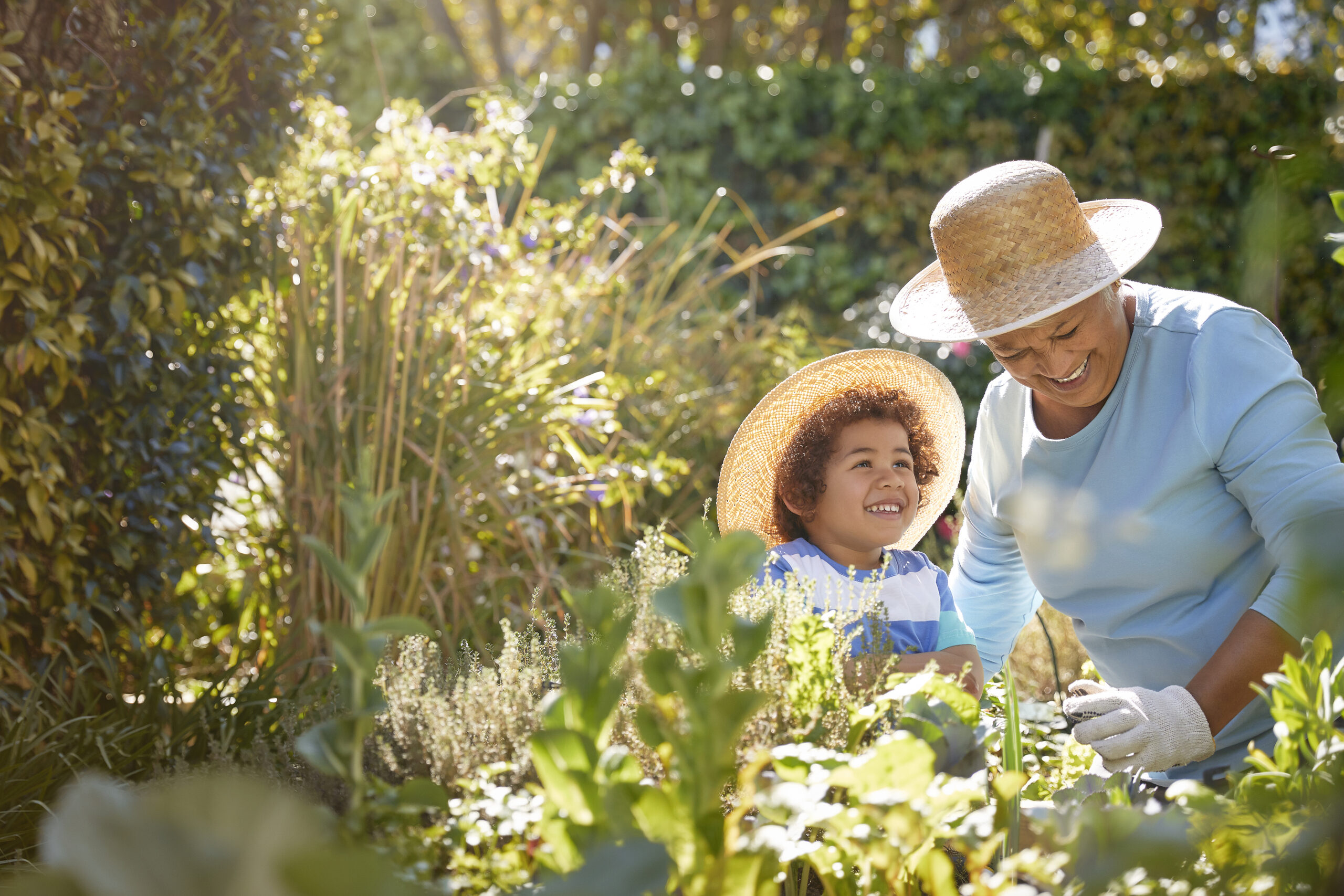An older woman and young child in sun hats smiling while gardening in a lush grassy place